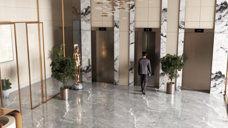 Interior view of a modern building lobby featuring three elevators with marble-clad walls. A man in a suit walks toward one of the elevators, while potted plants and a golden sculpture decorate the space. The floor is polished marble, and the design includes glass partitions and a contemporary ceiling light fixture.