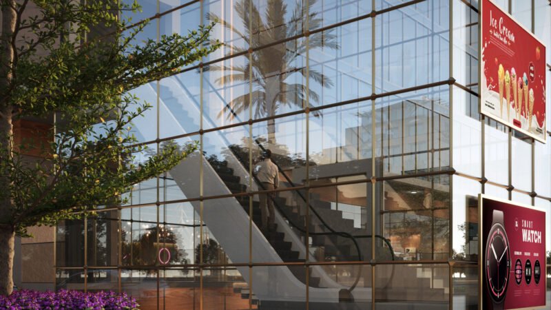 Exterior view of a modern glass-fronted commercial building showing an escalator inside. A person is seen ascending the escalator, with reflections of trees and palm trees on the glass panels. The building features outdoor signage advertising ice cream and smartwatches, and is surrounded by greenery and flowers.